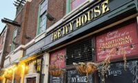 The Ivy House is a community-run pub in South East London. The image shows the front of the pub, with hanging baskets of flowers and chalkboards advertising the pub's hours.